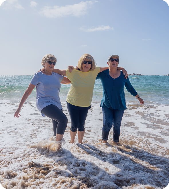 Three older women laughing as they walk in the shallows at the beach